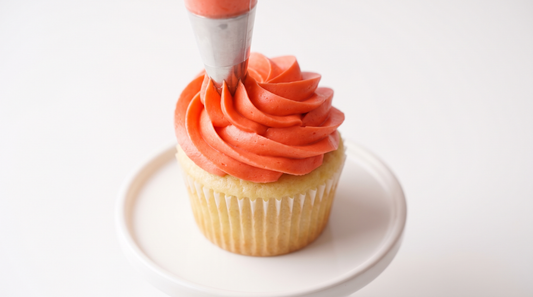 Vivid coral buttercream being piped onto a cupcake with crisp peaks
