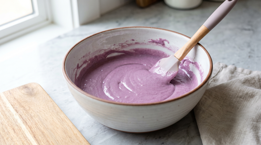 A bowl of vividly colored purple cupcake batter being folded with a spatula