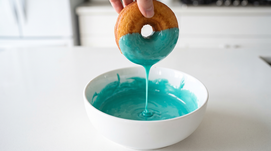 A donut being dipped into a bowl of thick glossy turquoise glaze